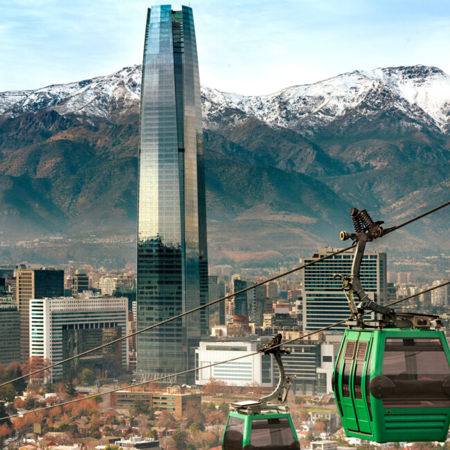 An image of a green gondola going up the hill in Santiago, Chile with the Costanera Tower and Andes mountains in the background